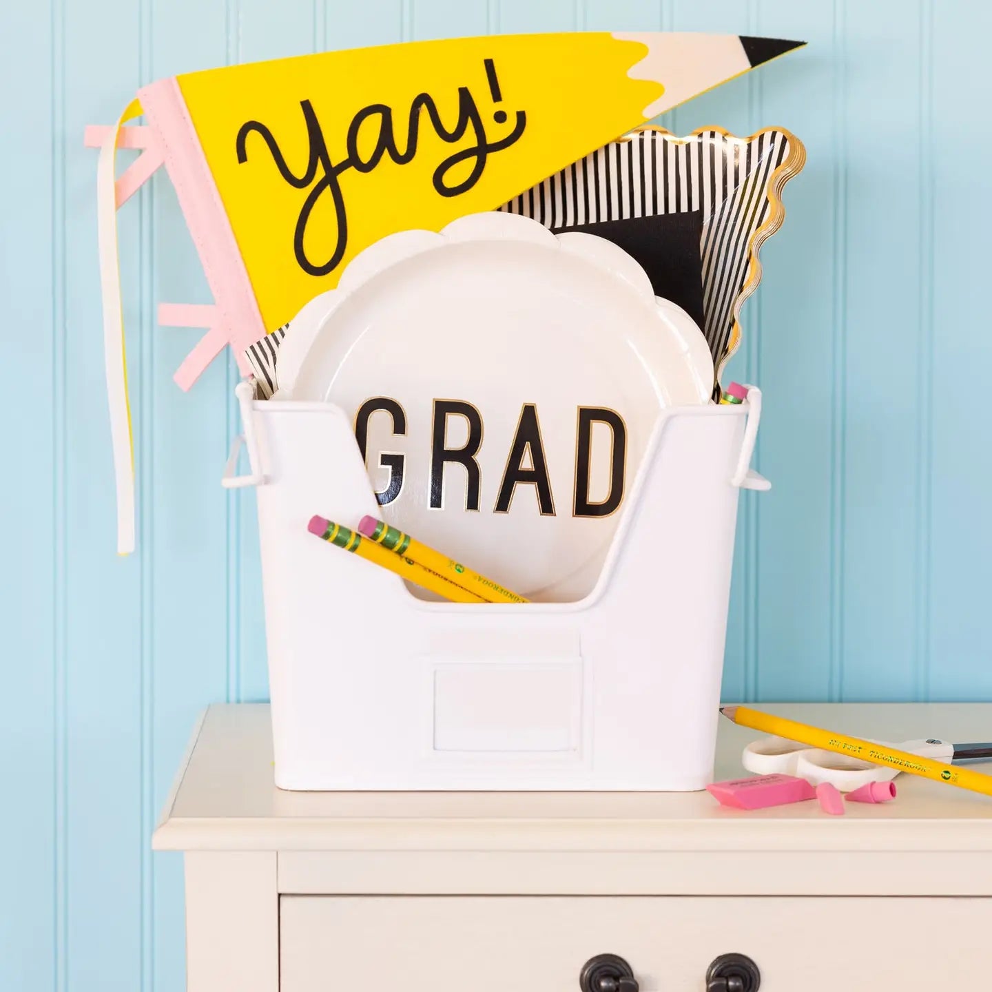 Graduation-themed decorative item with 'GRAD' bucket, colorful flags, and pencils on a light blue background.