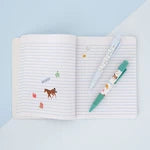 An open lined notebook on a pale blue background, decorated with small colorful stickers and featuring a green and white pen resting on the pages.