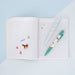 An open lined notebook on a pale blue background, decorated with small colorful stickers and featuring a green and white pen resting on the pages.