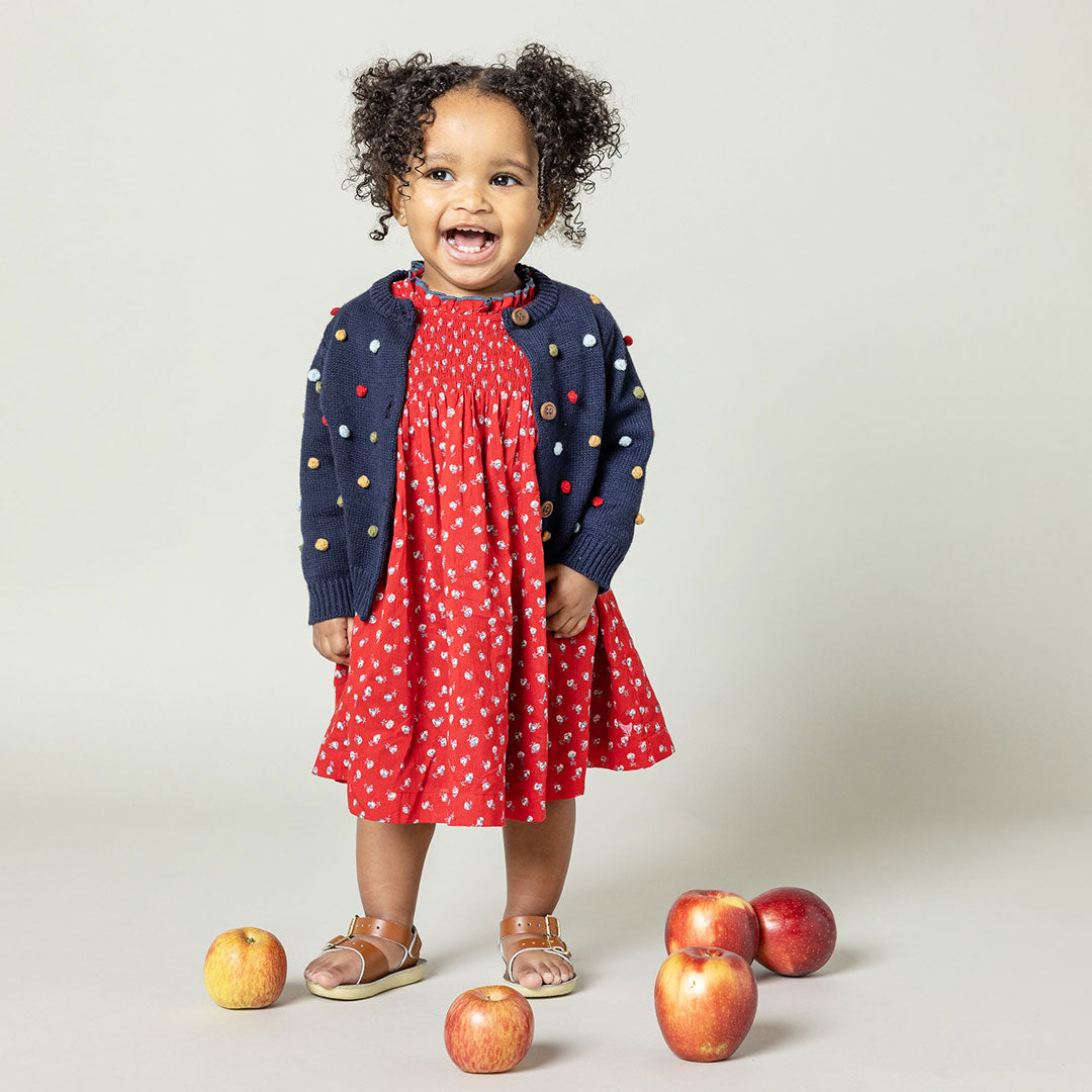 baby wearing red sleeveless dress with smocking at the chest and tiny white rose print and navy cardigan on top