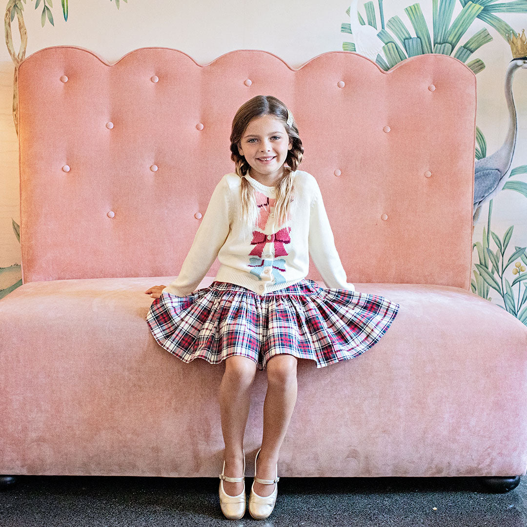 girl wearing cream colored cardigan with sparkle buttons and pink, red and blue knit bows down the middle over tartan plaid dress