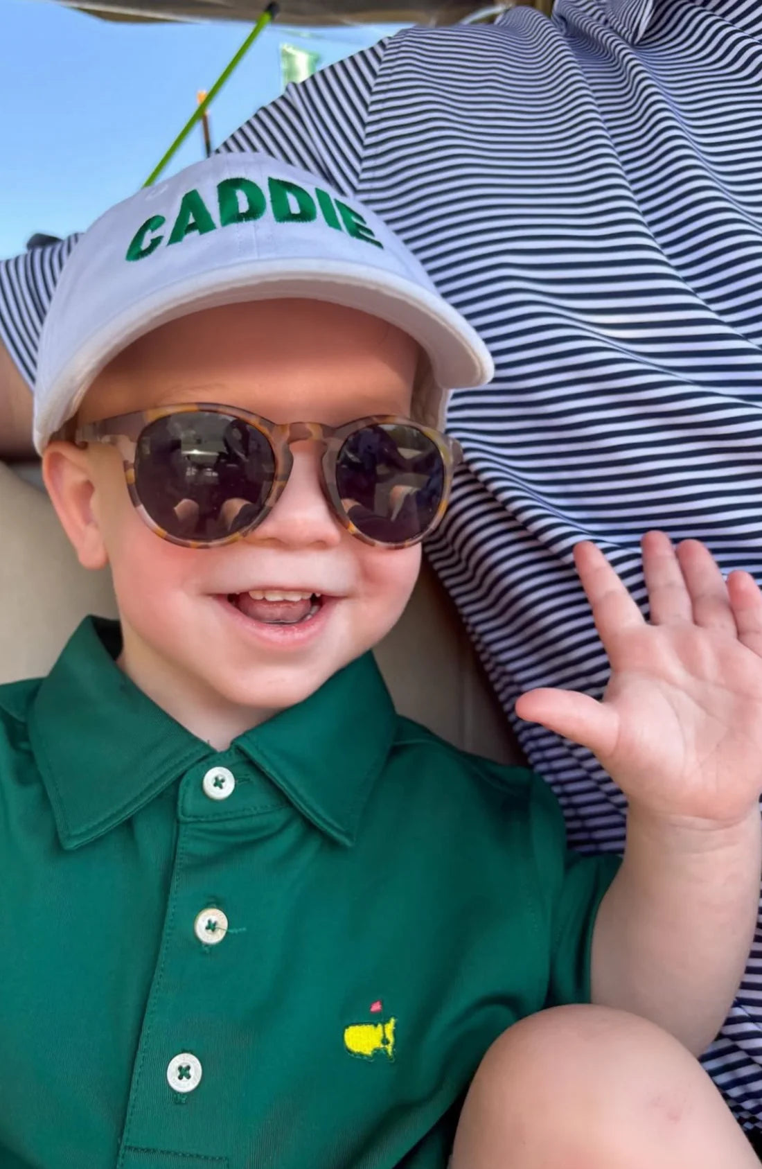 Child wearing a green shirt with a logo, sunglasses, and a cap with 'CADDIE' text.