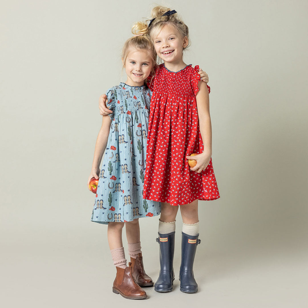 girl wearing light blue sleeveless dress with smocking and rodeo print including boots, cactus and cowboy hats