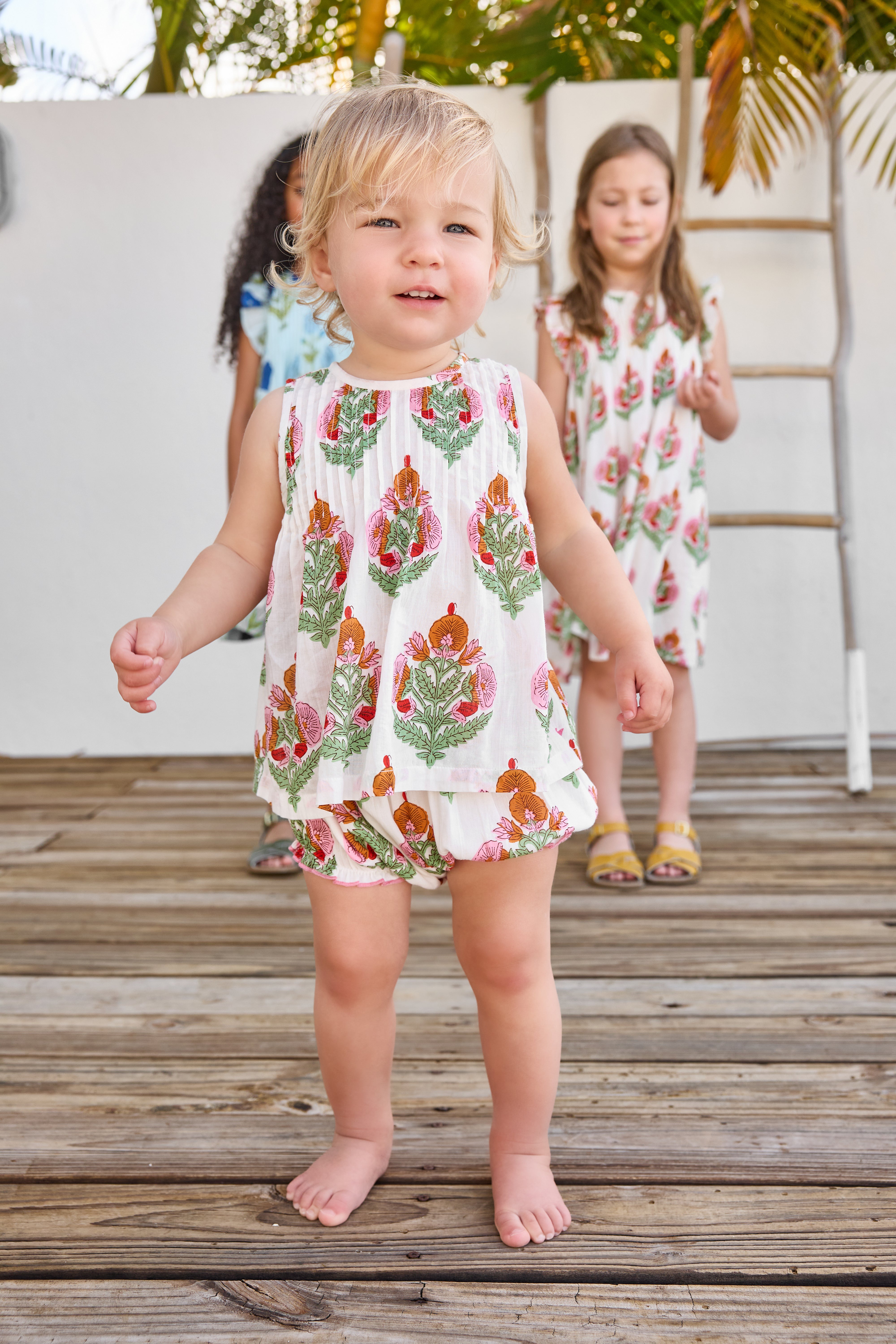 Two young girls in floral dresses standing on a wooden deck.