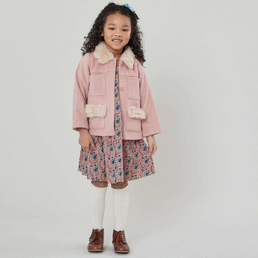girl wearing light pink pea coat with cream colored fur on pockets and collar with pink floral dress underneath