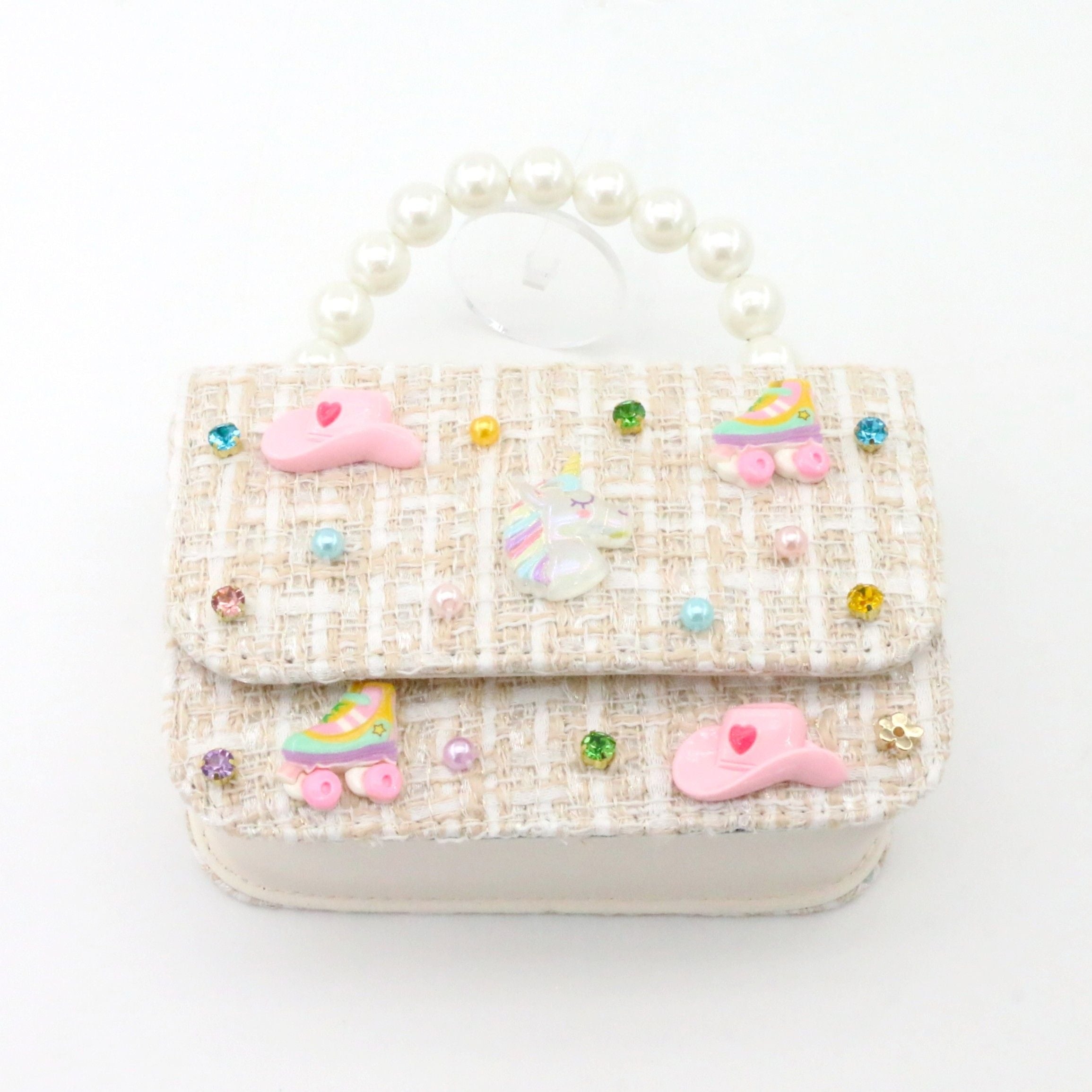 A small decorative handbag with a cream and beige tweed pattern, adorned with pink unicorn charms, pastel roller skates, and colorful rhinestones, featuring a pearl handle.