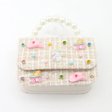 A small decorative handbag with a cream and beige tweed pattern, adorned with pink unicorn charms, pastel roller skates, and colorful rhinestones, featuring a pearl handle.