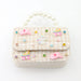 A small decorative handbag with a cream and beige tweed pattern, adorned with pink unicorn charms, pastel roller skates, and colorful rhinestones, featuring a pearl handle.