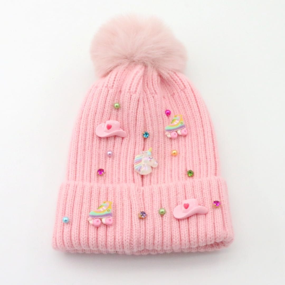 A light pink ribbed knit beanie hat with a fluffy pom-pom on top, decorated with pastel unicorn and roller skate embellishments and small rhinestone beads, shown on a plain white background.